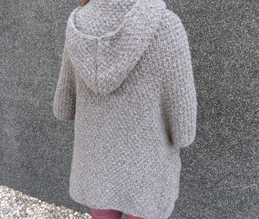 Rückansicht einer Person in einer hellgrauen, gestrickten Jacke mit Kapuze und dreiviertel Ärmeln vor einer dunklen Wand.