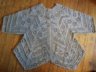 Gehäkelte Damenjacke in Grau-Beige mit Zacken- und Blütenmuster und drei Holzknöpfen, liegend auf Holzfußboden.