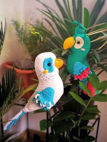 Gehäkelte Amigurumi-Vögel (Wellensittich und Papagei) zwischen Zimmerpflanzen.