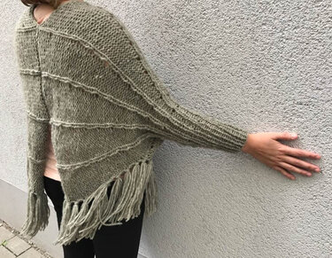 Rückansicht einer Person, die einen olivgrünen gestrickten Poncho mit horizontalen Streifen und Fransen trägt.