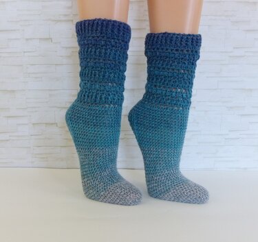 Paar gehäkelter Socken in Türkis-Grau an Beinmodellen vor heller Wand