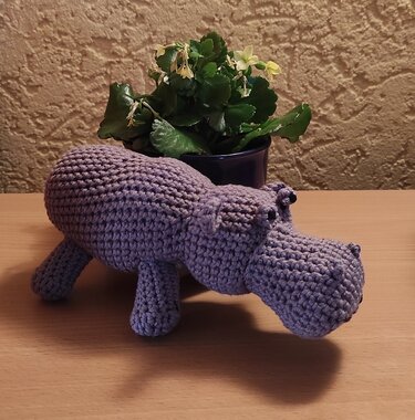 Handgehäkeltes lila Nilpferd-Amigurumi auf einem Holztisch vor einer Topfpflanze.