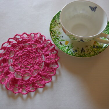 Fuchsiafarbener, gehäkelter Untersetzer neben einer Teetasse.