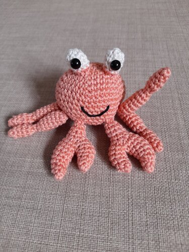 Gehäkelte zartrosa Krabbe mit weißen Augen und gesticktem Lächeln auf hellem Stoff