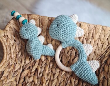 Zwei gehäkelte Dinosaurier in Mint und Creme, eine Rassel mit Holzring und eine kleine Dino-Figur am Perlenclip in einem geflochtenen Korb