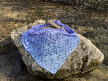 Gehäkeltes Dreieckstuch in blau-lila Farbverlauf mit Ajourmuster auf einem Felsen am Bach