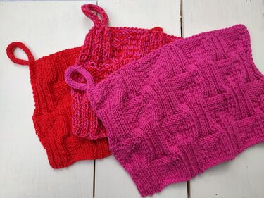 Drei gestrickte Topflappen in Rot und Pink mit Aufhängeschlaufen auf hellem Holzuntergrund