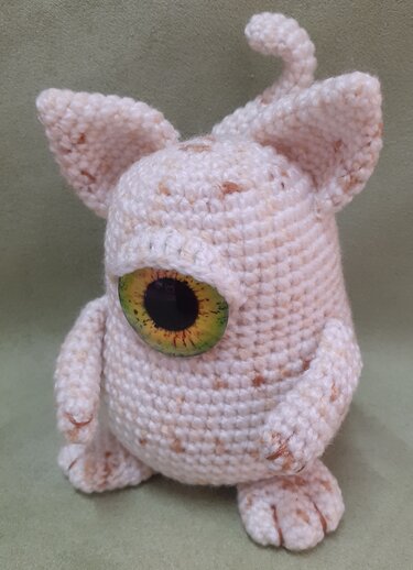 Cream-speckled crocheted amigurumi monster with one large green-and-yellow eye, pointy ears and a curled tail on a pale green background.