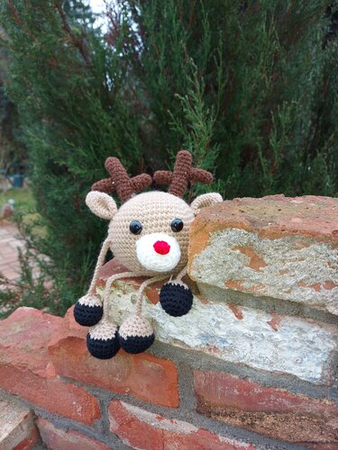 Beiges gehäkeltes Rentier-Amigurumi mit roter Nase und schwarzen Hufen sitzt auf einer Ziegelmauer vor grünen Sträuchern