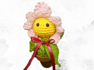 Gehäkelte Blume mit gelbem Gesicht, rosa Blütenblättern, grünen Blättern und roter Schleife im kleinen Tontopf