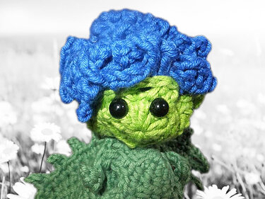 Gehäkelte grüne Figur mit blauem krausem Haar und schwarzen Knopfaugen vor Blumenwiese