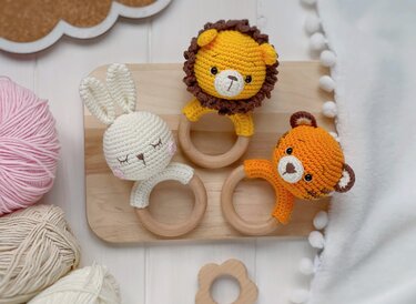 Three crocheted animal baby rattles with wooden rings: white bunny, yellow lion, and orange tiger on a wooden board with yarn and a blanket.