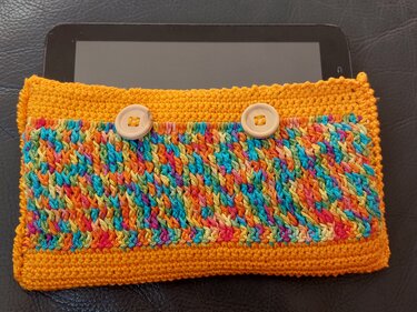 Gehäkelte orangefarbene Tablet-Hülle mit buntem Mittelstreifen und zwei hellen Holzknöpfen; Tablet teilweise sichtbar.