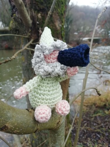 Gehäkelter Zwerg mit weißer Mütze, grauem Bart und blauem Horn auf einem Ast am Ufer