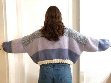 Rückenansicht einer Person in kurz geschnittener Oversize-Strickjacke mit pastellfarbenen Blockstreifen und ausgestreckten Armen