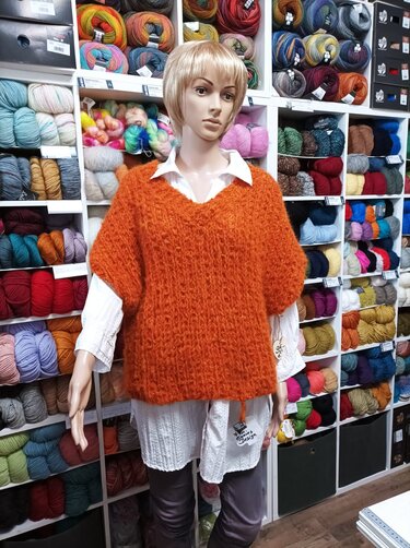 Mannequin mit orangefarbenem, locker gestricktem Poncho vor Regalen mit Wolle