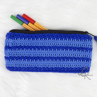 Gehäkelte Federtasche in Blautönen mit Streifenmuster und schwarzem Reißverschluss, aus dem bunte Stifte ragen.