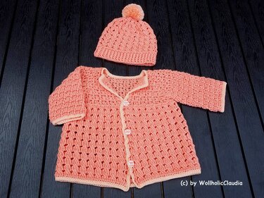 Gehäkelte Babyjacke mit Knöpfen und Korallen‑Bommelmütze auf dunklem Holz