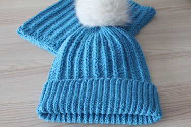 Blaues, geripptes Mützen- und Schalset mit weißem Bommel auf hellem Holz