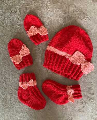Rotes und rosa gestricktes Baby-Set mit Schleifen: eine große Mütze, zwei kleine Mützen und ein Paar Socken auf grauem Untergrund