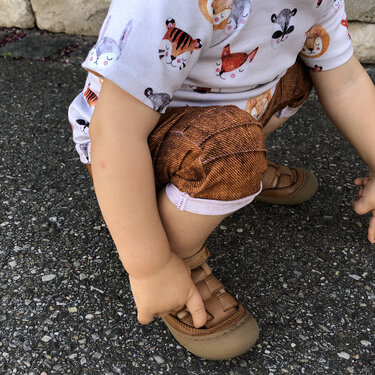 Nahaufnahme eines hockenden Kleinkinds, Fokus auf braune Baby-Shorts mit Umschlag und sichtbarer Naht; Hände greifen zur Sandale.