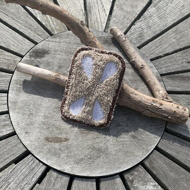 Beiges, rechteckiges ITH‑Stickmotiv mit weißem X und braunem Rand auf Holzuntergrund