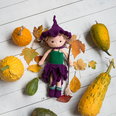 Gehäkelte Amigurumi-Hexe mit lila Hexenhut und kleinem Besen, umgeben von Herbstblättern und Zierkürbissen auf weißem Holzboden.