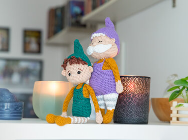Zwei gehäkelte Amigurumi-Wichtel in grüner und violetter Kleidung mit Kerzen im Hintergrund