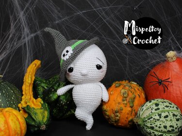 White crocheted ghost amigurumi wearing a grey witch hat with skull accent, shown with pumpkins, gourds and decorative spiderweb backdrop.