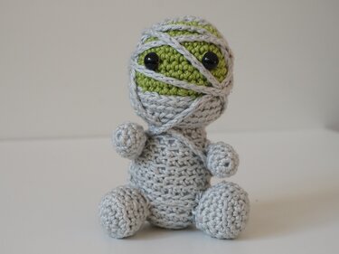 Gehäkelte sitzende Amigurumi
‑Mumie mit grünem Kopf, hellgrauen Bandagen und schwarzen Knopfaugen vor neutralem Hintergrund