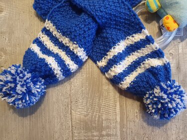 Gestrickter blau-weißer Babyschal mit zwei Bommeln auf Holzuntergrund