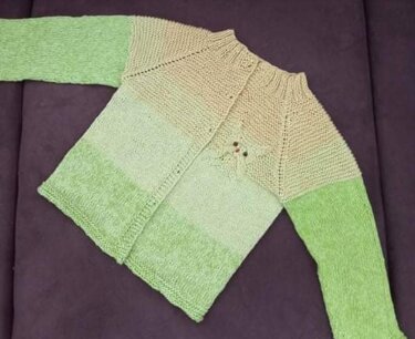 Flach liegende gestrickte Kinderjacke mit offenem Vorderteil, Raglan und Farbverlauf von Beige zu Grün auf dunklem Untergrund.