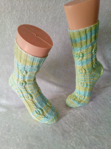 Ein Paar gestrickte Socken in hellgrün und blau gestreift mit Zopf- und Noppenmuster auf Fußmodellen vor hellem Hintergrund.