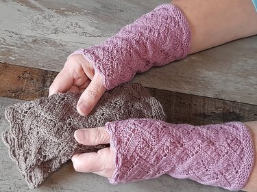 Zwei Hände mit gestrickten, fingerlosen Armstulpen: eine in Rosa am Handgelenk, die andere in Braun daneben liegend, beide mit Lochmuster.