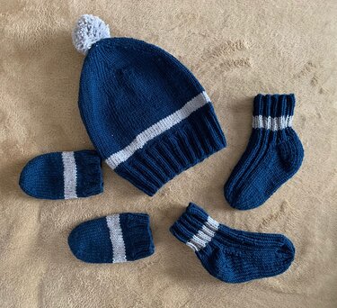 Gestricktes Babyset mit Mütze und Bommel, ein Paar Söckchen und ein Paar Fäustlinge in Marineblau mit hellgrauen Streifen auf beigem Untergrund.