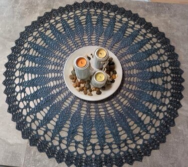 Rundes gehäkeltes Deckchen in Blau- und Schwarztönen mit Spitzenmuster und gezacktem Rand. In der Mitte ein Teller mit drei Kerzen und Kieselsteinen.