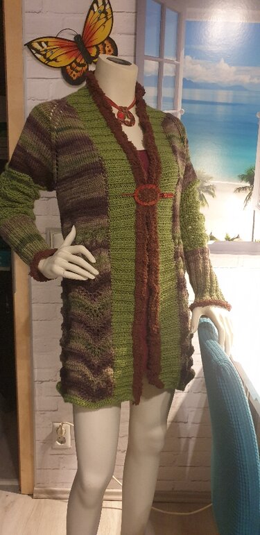 Mannequin mit langer, offen getragener Strickjacke in Grün- und Brauntönen, gemustert, mit flauschigen Rändern und runder Zierschließe