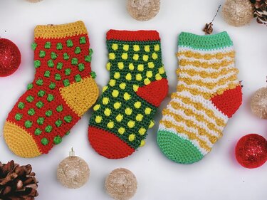 Drei kleine gehäkelte Weihnachtsstrümpfe in Rot, Grün und Gelb mit Noppenmuster, umgeben von Kugeln und Tannenzapfen.