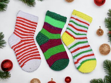 Three striped crochet Christmas stockings with varied colorways, placed beside pine sprigs and round ornaments.