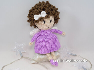 Gehäkeltes Amigurumi-Engelchen mit lila Kleid, braunen Locken und weißen Flügeln, auf hellem Untergrund sitzend