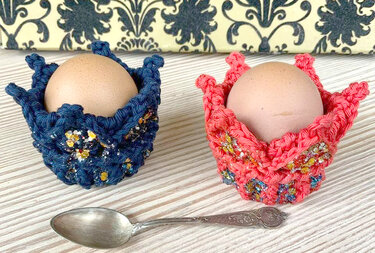Zwei gehäkelte Eierwärmer in Blau und Koralle mit Glitzerdetails, jeweils ein braunes Ei; ein Silberlöffel liegt auf einem hellen Holztisch.