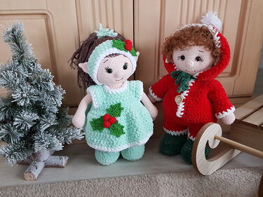 Zwei gehäkelte Amigurumi-Puppen in mintfarbenem Weihnachtskleid und rotem Kapuzenmantel neben kleinem Kunstbaum