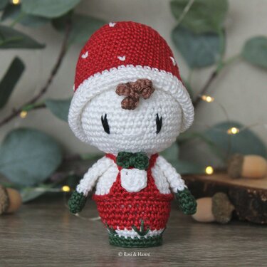 Gehäkelte Amigurumi-Glückspilz-Figur mit roter Mütze, roter Latzhose und grüner Fliege auf Holzfläche.