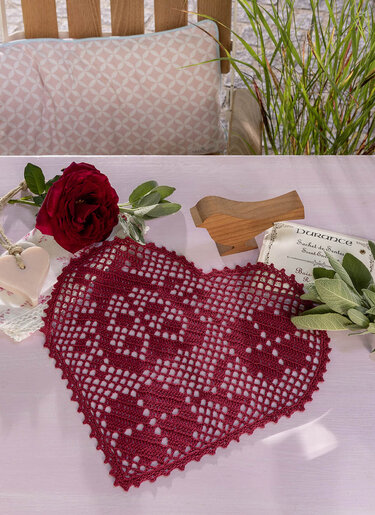 Gehäkeltes herzförmiges Deckchen dunkelrot mit Rose, Salbei und Holzvogel auf hellem Tisch
