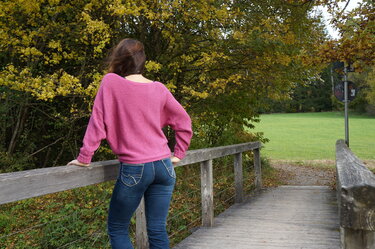 Rückenansicht eines weit geschnittenen rosa Strickpullovers mit Bündchen, getragen von Frau auf einer Holzbrücke; Jeans und herbstliche Bäume im Hintergrund