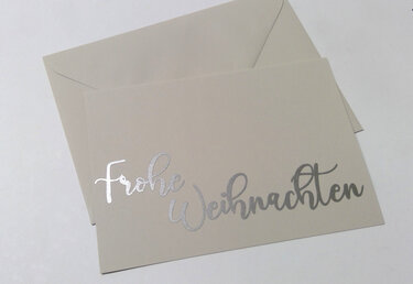 Beigefarbene Karte mit silbernem Schriftzug „Frohe Weihnachten“ und passendem Umschlag