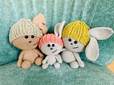 Nahaufnahme von drei gehäkelten Amigurumi-Hasen: ein beiger Hase, ein kleiner grauer Hase mit rosa Mütze und ein größerer grauer Hase mit gelber Mütze und langen Ohren.