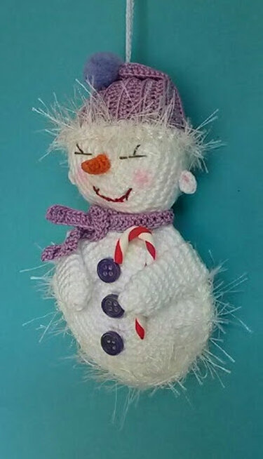 Gehäkelter weißer Amigurumi-Schneemann mit lila Mütze und Schal, hält Zuckerstange