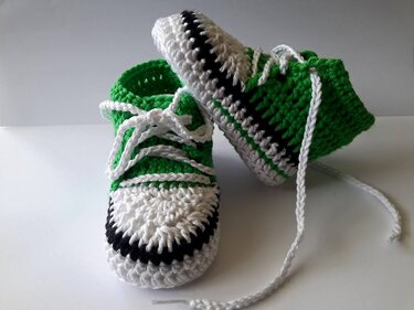 Handgehäkelte Babyschuhe im Sneaker‑Stil in Grün, Weiß und Schwarz mit weißen Schnürsenkeln.