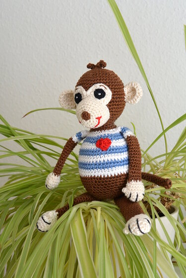 Gehäkelter brauner Amigurumi-Affe mit blau-weiß gestreiftem Shirt und rotem Herz auf grüner Pflanze